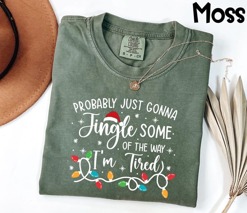 Puede incluir: Una camiseta verde musgo con el texto "Probably Just Gonna Jingle Some of the Way I'm Tired" en blanco. El dise&ntilde;o incluye un gorro de Pap&aacute; Noel y luces de cadena. Un collar dorado est&aacute; drapeado sobre la camisa.