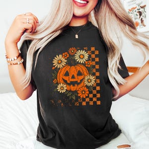 Puede incluir: Camiseta negra con un dise&ntilde;o de Halloween de estilo vintage. El gr&aacute;fico incluye una calabaza naranja rodeada de flores naranjas y amarillas, con un patr&oacute;n de cuadros en el lado derecho. El dise&ntilde;o tiene un aspecto envejecido.