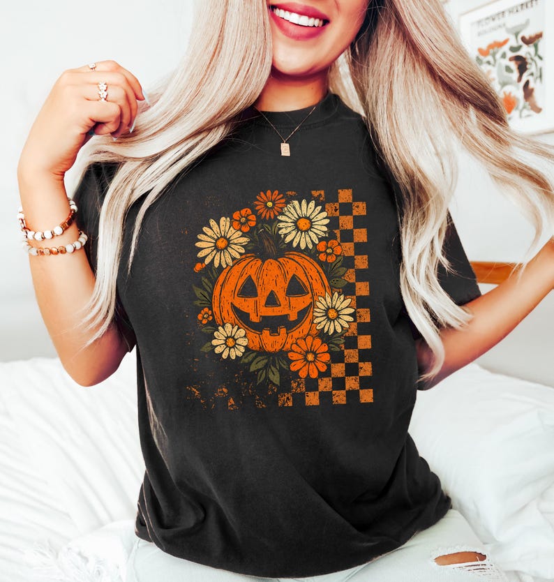 Puede incluir: Camiseta negra con un dise&ntilde;o de Halloween de estilo vintage. El gr&aacute;fico incluye una calabaza naranja rodeada de flores naranjas y amarillas, con un patr&oacute;n de cuadros en el lado derecho. El dise&ntilde;o tiene un aspecto envejecido.