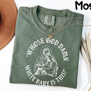 May include: Moss green t-shirt with the text "WHOSE GOD DAMN WHITE BABY IS THIS?" in a circular design around an illustration of a religious family. The shirt has a crew neck and short sleeves.