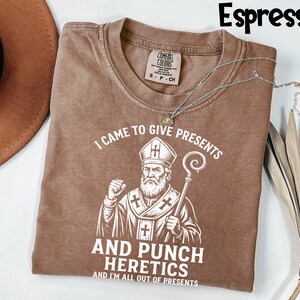 May include: Espresso-coloured t-shirt with the text "I CAME TO GIVE PRESENTS AND PUNCH HERETICS AND I'M ALL OUT OF PRESENTS" and a graphic of a religious figure. A silver necklace is draped across the shirt.