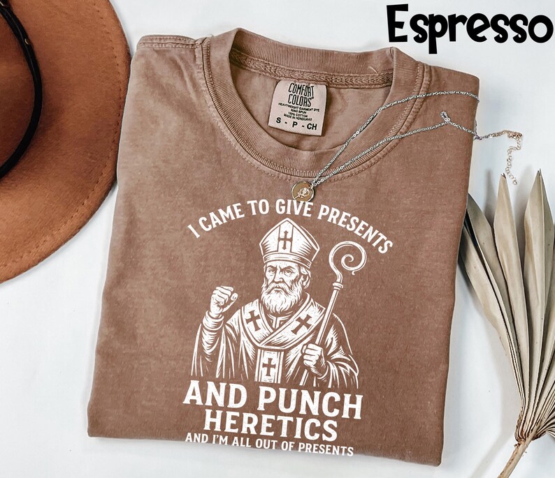 May include: Espresso-coloured t-shirt with the text "I CAME TO GIVE PRESENTS AND PUNCH HERETICS AND I'M ALL OUT OF PRESENTS" and a graphic of a religious figure. A silver necklace is draped across the shirt.
