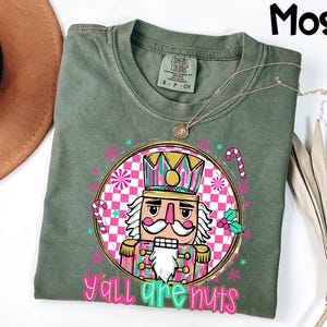 May include: A moss green t-shirt featuring a colorful graphic of a nutcracker with the text "Y'all are nuts." The design includes pink, teal, and gold accents, with candy canes and snowflakes. The shirt has a crew neck and short sleeves.