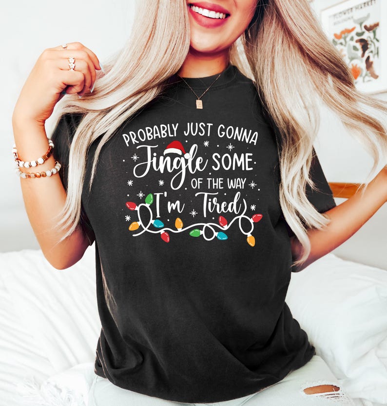 Puede incluir: Camiseta negra con la frase "Probably just gonna jingle some of the way, I'm tired." El dise&ntilde;o incluye un gorro de Pap&aacute; Noel, copos de nieve y una guirnalda de luces. Una prenda casual y festiva.