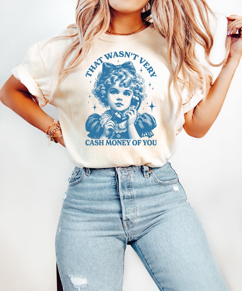 Puede incluir: Una camiseta blanca con una ilustraci&oacute;n azul de una ni&ntilde;a al tel&eacute;fono. El texto en la camiseta dice "That wasn't very cash money of you."