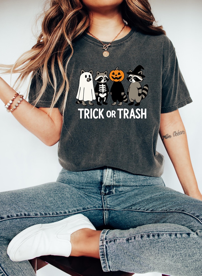 May include: Dark gray t-shirt with a Halloween-themed graphic. The design features four cartoon raccoons dressed as a ghost, skeleton, pumpkin, and witch, with the words "TRICK OR TRASH" in white block letters.