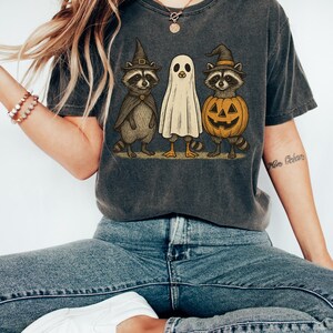 May include: Dark gray t-shirt featuring a Halloween-themed graphic. The design includes a raccoon in a witch hat and cape, a ghost, and a raccoon inside a jack-o'-lantern. The shirt is worn by a person wearing blue jeans and white sneakers.