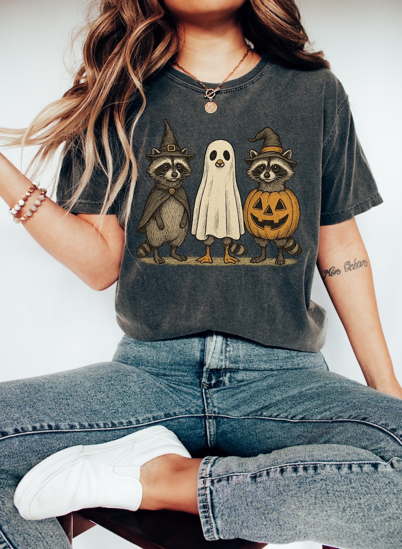 May include: Dark gray t-shirt featuring a Halloween-themed graphic. The design includes a raccoon in a witch hat and cape, a ghost, and a raccoon inside a jack-o'-lantern. The shirt is worn by a person wearing blue jeans and white sneakers.
