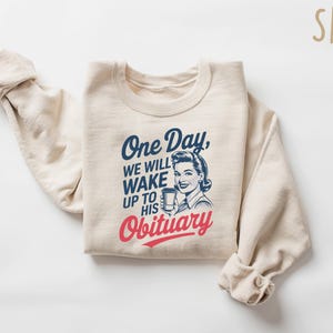 May include: A beige sweatshirt with the text "One Day, We Will Wake Up To His Obituary" in blue and red lettering. A retro-style illustration of a woman holding a cup is above the text.