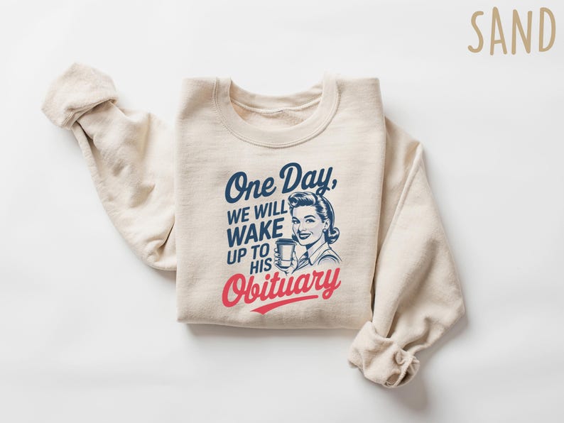 May include: A beige sweatshirt with the text "One Day, We Will Wake Up To His Obituary" in blue and red lettering. A retro-style illustration of a woman holding a cup is above the text.