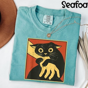 May include: Seafoam green t-shirt with a graphic of a black cat biting a hand. The cat is in a red and yellow square. A gold necklace with a pendant is on the shirt. The shirt is labeled "Comfort Colors".