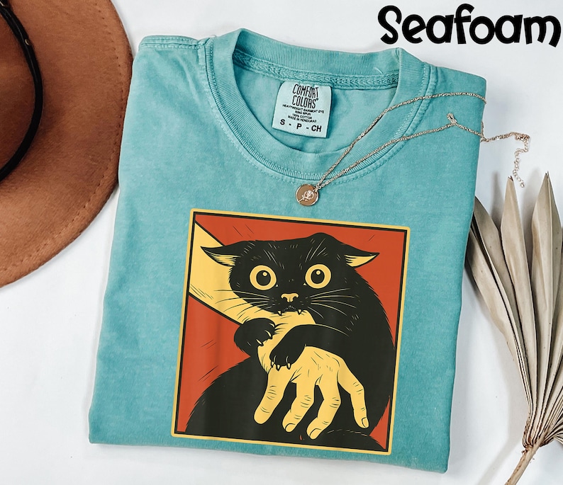 May include: Seafoam green t-shirt with a graphic of a black cat biting a hand. The cat is in a red and yellow square. A gold necklace with a pendant is on the shirt. The shirt is labeled "Comfort Colors".