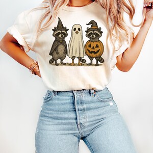 May include: Cream-colored t-shirt featuring a Halloween-themed graphic. The design includes three cartoon characters: two raccoons in witch hats and capes, and a ghost. One raccoon holds a jack-o'-lantern. The shirt is paired with light blue jeans.