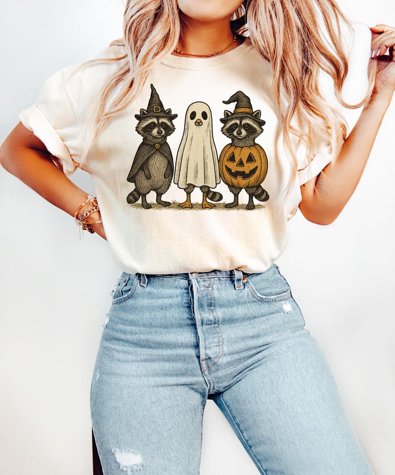 May include: Cream-colored t-shirt featuring a Halloween-themed graphic. The design includes three cartoon characters: two raccoons in witch hats and capes, and a ghost. One raccoon holds a jack-o'-lantern. The shirt is paired with light blue jeans.