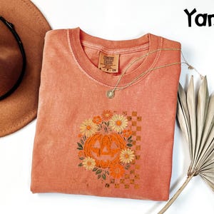 Puede incluir: Una camiseta Comfort Colors de color coral con cuello redondo, con un gr&aacute;fico de calabaza rodeada de flores. El dise&ntilde;o de la calabaza es naranja y amarillo, sobre un fondo a cuadros. Un collar dorado y un sombrero marr&oacute;n tambi&eacute;n est&aacute;n en la imagen.
