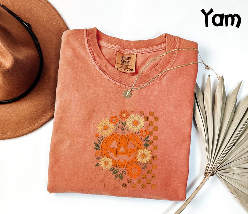 Puede incluir: Una camiseta Comfort Colors de color coral con cuello redondo, con un gr&aacute;fico de calabaza rodeada de flores. El dise&ntilde;o de la calabaza es naranja y amarillo, sobre un fondo a cuadros. Un collar dorado y un sombrero marr&oacute;n tambi&eacute;n est&aacute;n en la imagen.