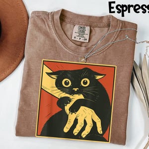 May include: Espresso-colored t-shirt with a graphic of a black cat biting a yellow hand, set against a red background. The shirt has a crew neck and a Comfort Colors tag. A silver necklace is draped across the shirt.