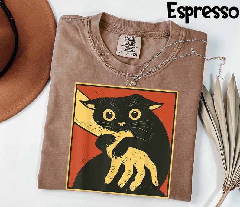 May include: Espresso-colored t-shirt with a graphic of a black cat biting a yellow hand, set against a red background. The shirt has a crew neck and a Comfort Colors tag. A silver necklace is draped across the shirt.