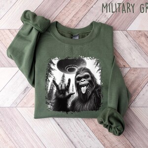 May include: A green sweatshirt with a black and white graphic print of a Bigfoot giving the 'I love you' hand sign with a UFO in the background. The text 'MILITARY GREEN' is printed in white on the top right corner of the image.