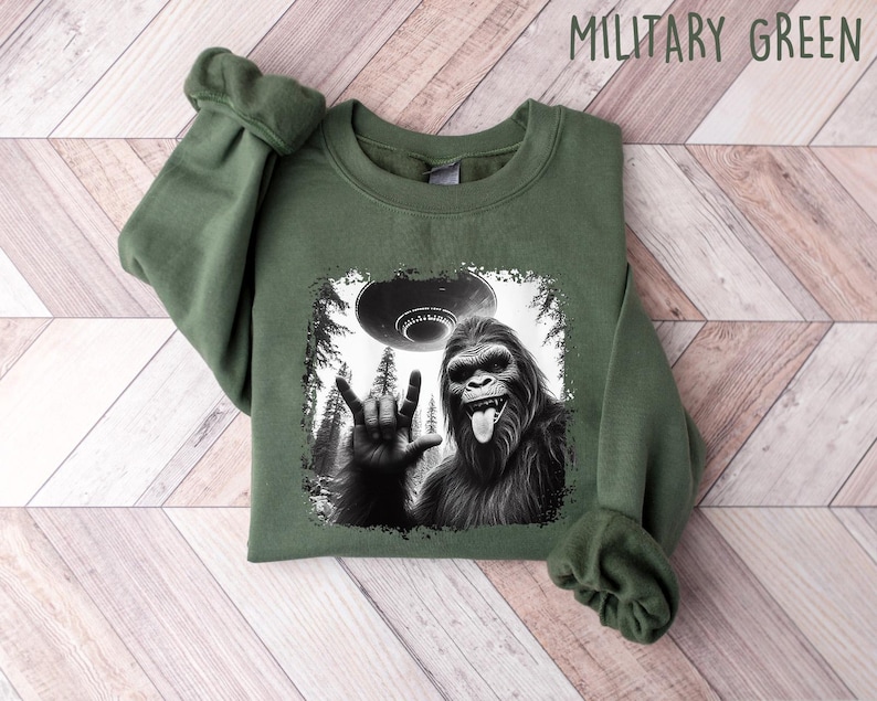 May include: A green sweatshirt with a black and white graphic print of a Bigfoot giving the 'I love you' hand sign with a UFO in the background. The text 'MILITARY GREEN' is printed in white on the top right corner of the image.