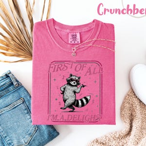 May include: A cerise t-shirt featuring a raccoon graphic with the text "FIRST OF ALL I'M A DELIGHT". The shirt is styled with blue jeans, white trainers, and a necklace. The word "Crunchberry" is written in pink script at the top of the image.
