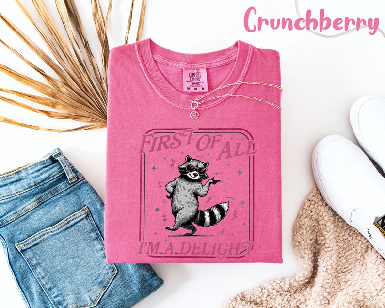 May include: A cerise t-shirt featuring a raccoon graphic with the text "FIRST OF ALL I'M A DELIGHT". The shirt is styled with blue jeans, white trainers, and a necklace. The word "Crunchberry" is written in pink script at the top of the image.