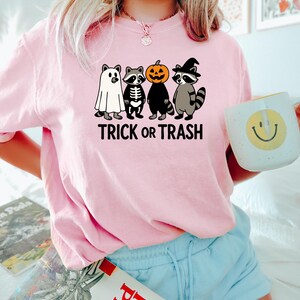 May include: A light pink sweatshirt featuring a graphic of four raccoons dressed in Halloween costumes: a ghost, a skeleton, a pumpkin, and a witch. The text "TRICK OR TRASH" is printed below the raccoons. The person is wearing blue shorts and holding a mug.
