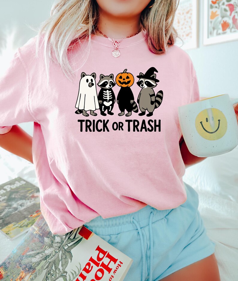 May include: A light pink sweatshirt featuring a graphic of four raccoons dressed in Halloween costumes: a ghost, a skeleton, a pumpkin, and a witch. The text "TRICK OR TRASH" is printed below the raccoons. The person is wearing blue shorts and holding a mug.