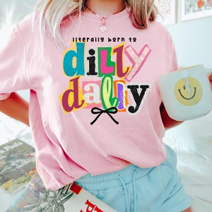 以下が含まれることがあります： カラフルな文字で「literally born to dilly dally」と書かれたピンク色のスウェットシャツ。黄色いスマイリーフェイスの白いマグカップが手に持たれています。青いショートパンツを着用しています。