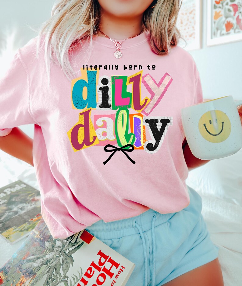 以下が含まれることがあります： カラフルな文字で「literally born to dilly dally」と書かれたピンク色のスウェットシャツ。黄色いスマイリーフェイスの白いマグカップが手に持たれています。青いショートパンツを着用しています。