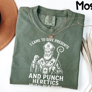 May include: A moss green t-shirt featuring a graphic of a religious figure with a staff. The text on the shirt reads "I CAME TO GIVE PRESENTS AND PUNCH HERETICS AND I'M ALL OUT OF PRESENTS". A gold necklace is draped across the shirt.