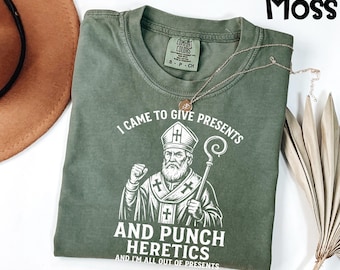 May include: Moss green t-shirt with a graphic of a religious figure holding a staff. The text on the shirt reads "I CAME TO GIVE PRESENTS AND PUNCH HERETICS AND I'M ALL OUT OF PRESENTS". A gold necklace is draped across the shirt.