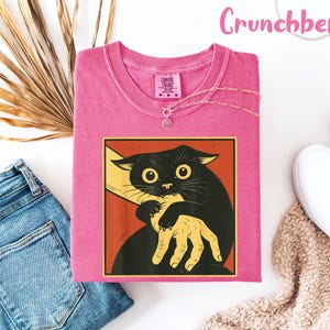 May include: A pink t-shirt with a graphic of a black cat biting a hand. The cat has large yellow eyes and is set against a red and yellow background. The text "Crunchberry" is in pink script at the top. A silver necklace and white shoes are also visible.