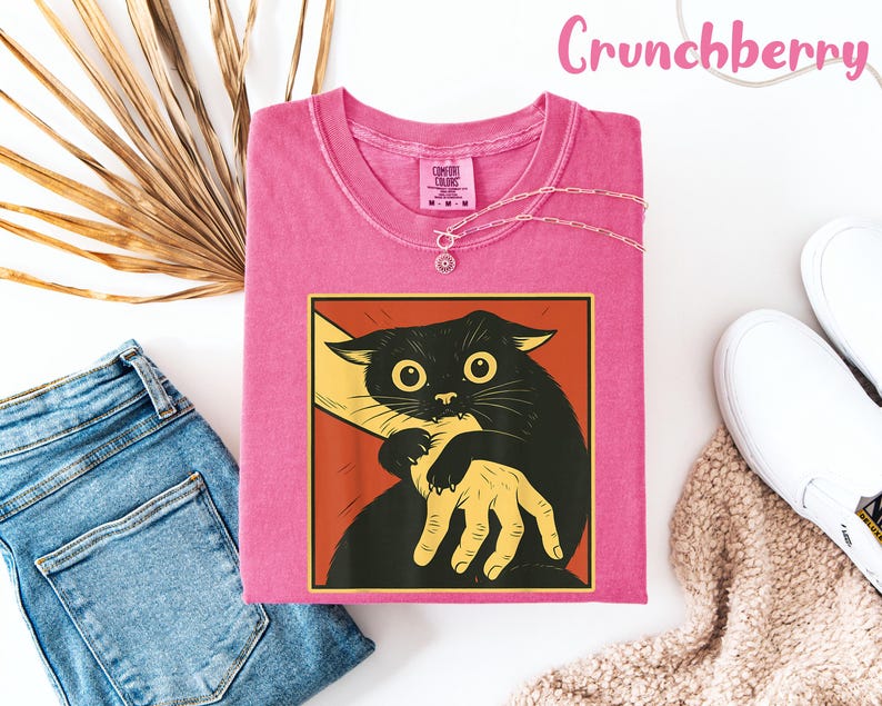 May include: A pink t-shirt with a graphic of a black cat biting a hand. The cat has large yellow eyes and is set against a red and yellow background. The text "Crunchberry" is in pink script at the top. A silver necklace and white shoes are also visible.