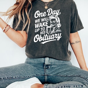 May include: A dark gray t-shirt with a white graphic print of a woman holding a coffee cup. The text on the shirt reads "One Day, We Will Wake Up To His Obituary."