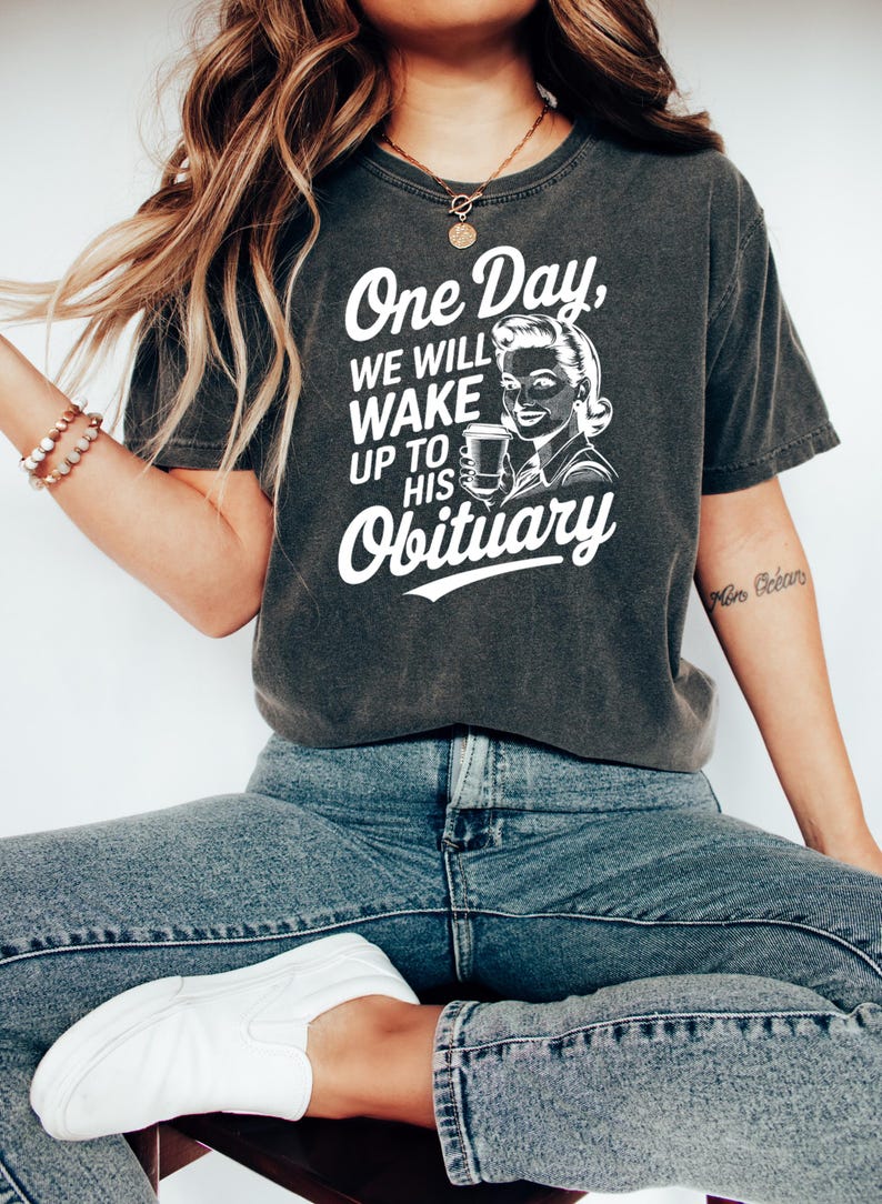 May include: A dark gray t-shirt with a white graphic print of a woman holding a coffee cup. The text on the shirt reads "One Day, We Will Wake Up To His Obituary."