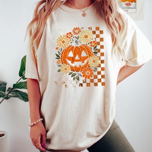 Puede incluir: Camiseta color crema con un dise&ntilde;o tem&aacute;tico de Halloween. El dise&ntilde;o incluye una calabaza naranja rodeada de flores naranjas y amarillas, sobre un fondo a cuadros. La camiseta es de corte holgado.