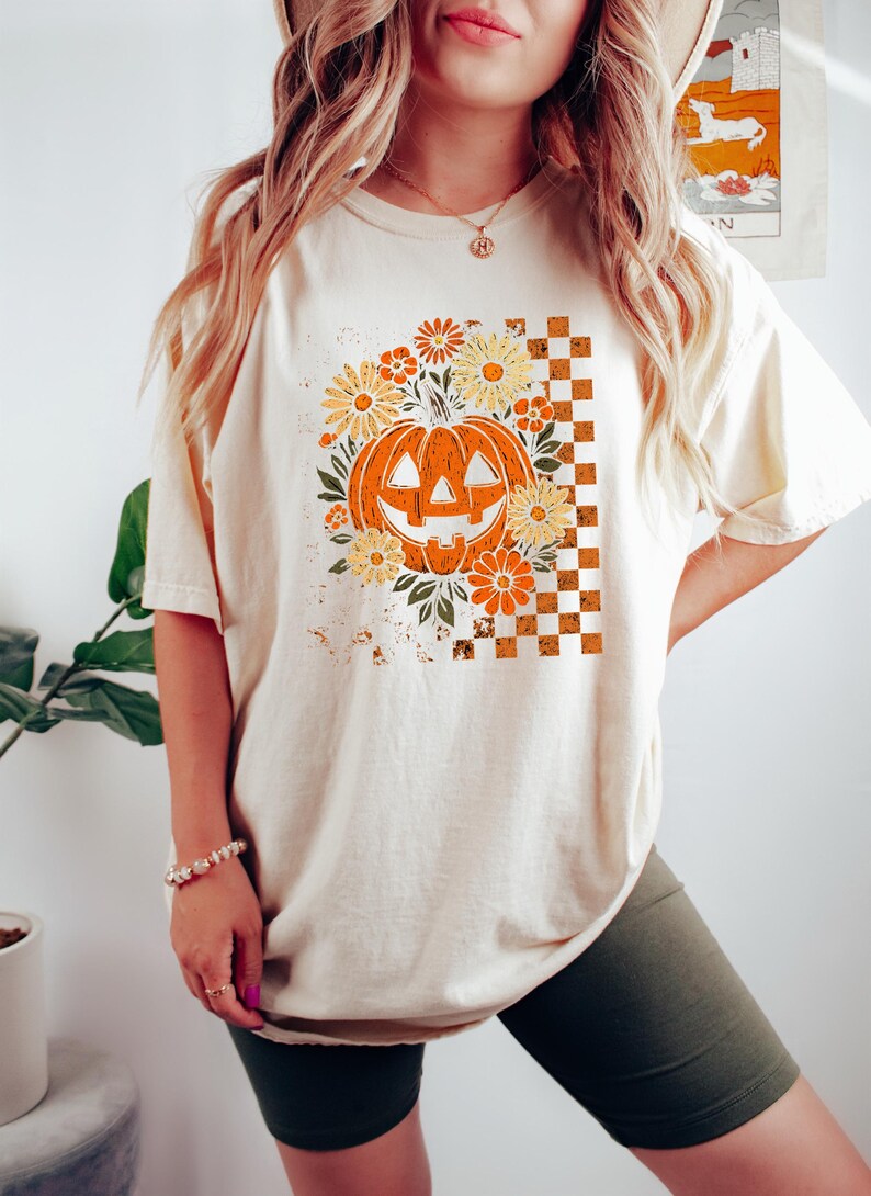 Puede incluir: Camiseta color crema con un dise&ntilde;o tem&aacute;tico de Halloween. El dise&ntilde;o incluye una calabaza naranja rodeada de flores naranjas y amarillas, sobre un fondo a cuadros. La camiseta es de corte holgado.
