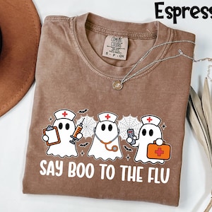 May include: Espresso-colored t-shirt featuring a graphic of three cartoon ghosts dressed as nurses, with medical tools and the text "SAY BOO TO THE FLU". The shirt has a crew neck and is made of a soft material. A silver necklace is draped across the shirt.