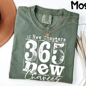 May include: A moss green t-shirt with the text "12 New Chapters 365 New Chances" in white. A gold necklace with a circular pendant rests on the shirt. The shirt is folded, and a brown hat is visible in the background.