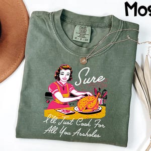 May include: Moss green t-shirt with a retro-style graphic of a woman cooking a turkey. The text on the shirt reads "Sure, I'll Just Cook For All You Assholes." A gold necklace is draped across the shirt.