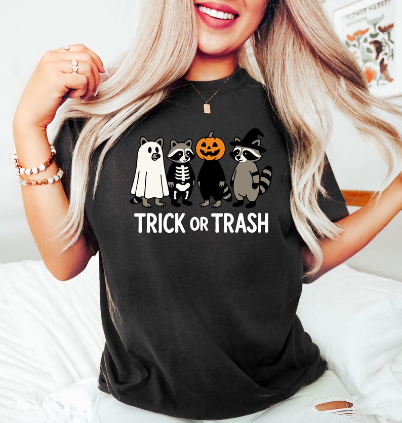 May include: Black t-shirt featuring a Halloween-themed graphic. The design includes four cartoon raccoons dressed in costumes: a ghost, a skeleton, a jack-o'-lantern, and a witch. The text "TRICK OR TRASH" is printed below the characters.