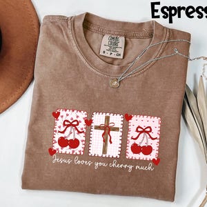 May include: Espresso-colored t-shirt with a graphic design featuring cherries, a cross, and the text "Jesus loves you cherry much." The shirt has a crew neck and short sleeves. A silver necklace with a pendant is draped on the shirt.