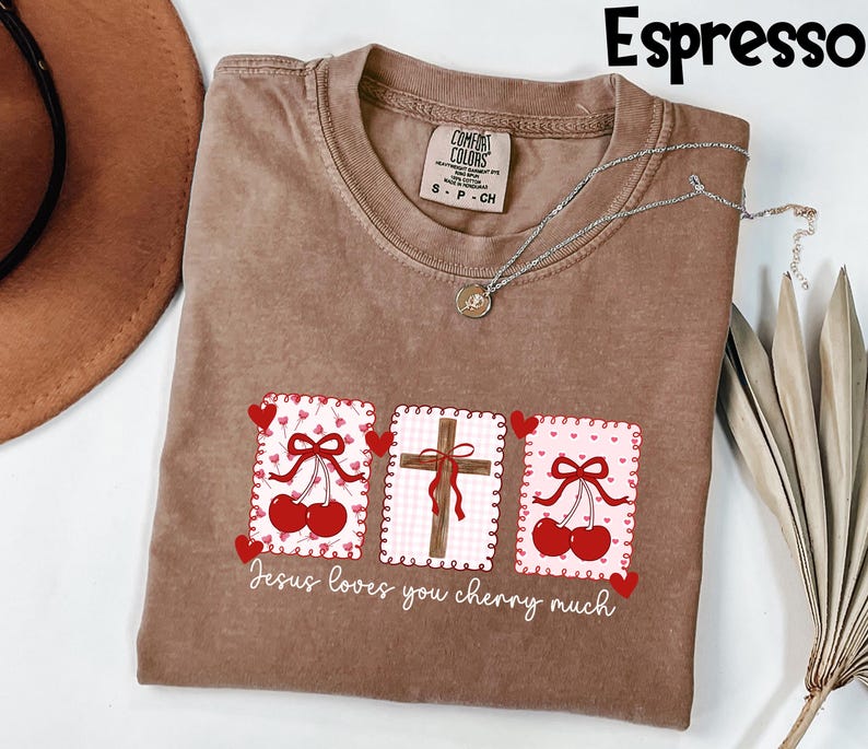 May include: Espresso-colored t-shirt with a graphic design featuring cherries, a cross, and the text "Jesus loves you cherry much." The shirt has a crew neck and short sleeves. A silver necklace with a pendant is draped on the shirt.
