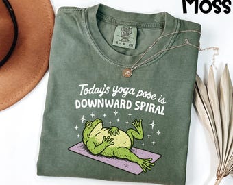 May include: Moss green t-shirt with the text "Today's yoga pose is DOWNWARD SPIRAL" and a cartoon frog on a yoga mat. The frog is green with a yellow belly. A gold necklace is draped across the shirt.