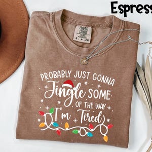 Puede incluir: Camiseta color espresso con el texto blanco "PROBABLY JUST GONNA JINGLE SOME OF THE WAY I'M TIRED". El dise&ntilde;o incluye un gorro de Pap&aacute; Noel, copos de nieve y una guirnalda de luces navide&ntilde;as de colores. Un collar plateado con un colgante est&aacute; sobre la camiseta.