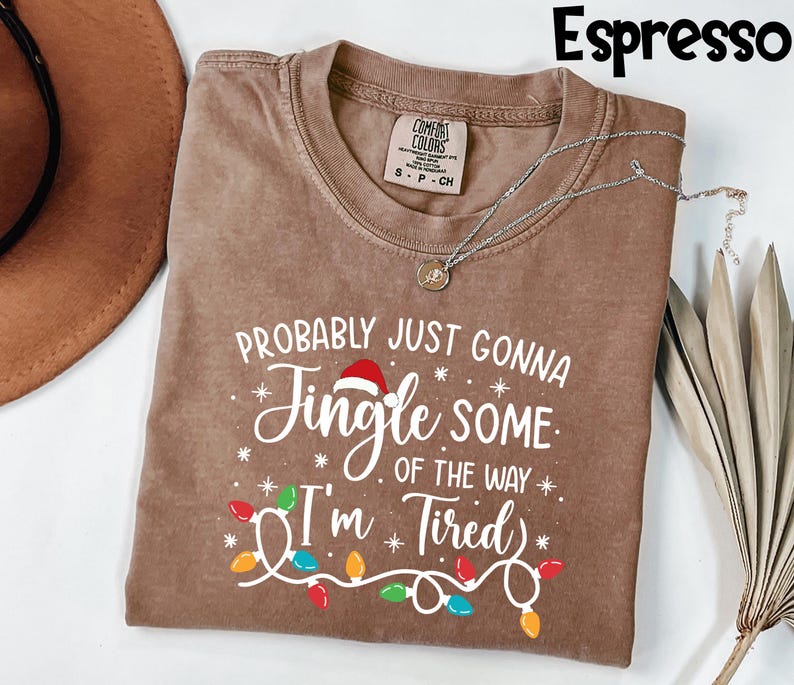 Puede incluir: Camiseta color espresso con el texto blanco "PROBABLY JUST GONNA JINGLE SOME OF THE WAY I'M TIRED". El dise&ntilde;o incluye un gorro de Pap&aacute; Noel, copos de nieve y una guirnalda de luces navide&ntilde;as de colores. Un collar plateado con un colgante est&aacute; sobre la camiseta.