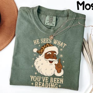 May include: A moss green t-shirt with a graphic of Santa Claus wearing a leopard print hat and the text "HE SEES WHAT YOU'VE BEEN READING". The shirt has a crew neck and short sleeves.