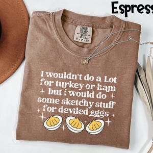 May include: Espresso-colored t-shirt with the text "I wouldn't do a Lot for turkey or ham + but I would do + some sketchy stuff + for deviled eggs +" and a graphic of deviled eggs. A silver necklace is draped across the shirt.