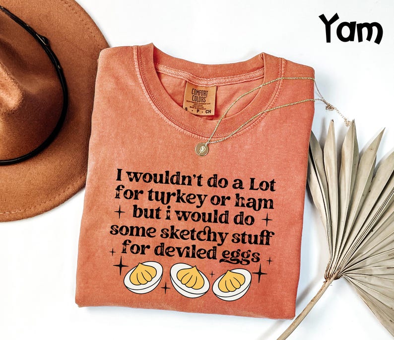 May include: A coral-colored t-shirt with the text "I wouldn't do a Lot for turkey or ham + but I would do + some sketchy stuff for deviled eggs +" and a graphic of deviled eggs. A gold necklace is draped across the shirt.